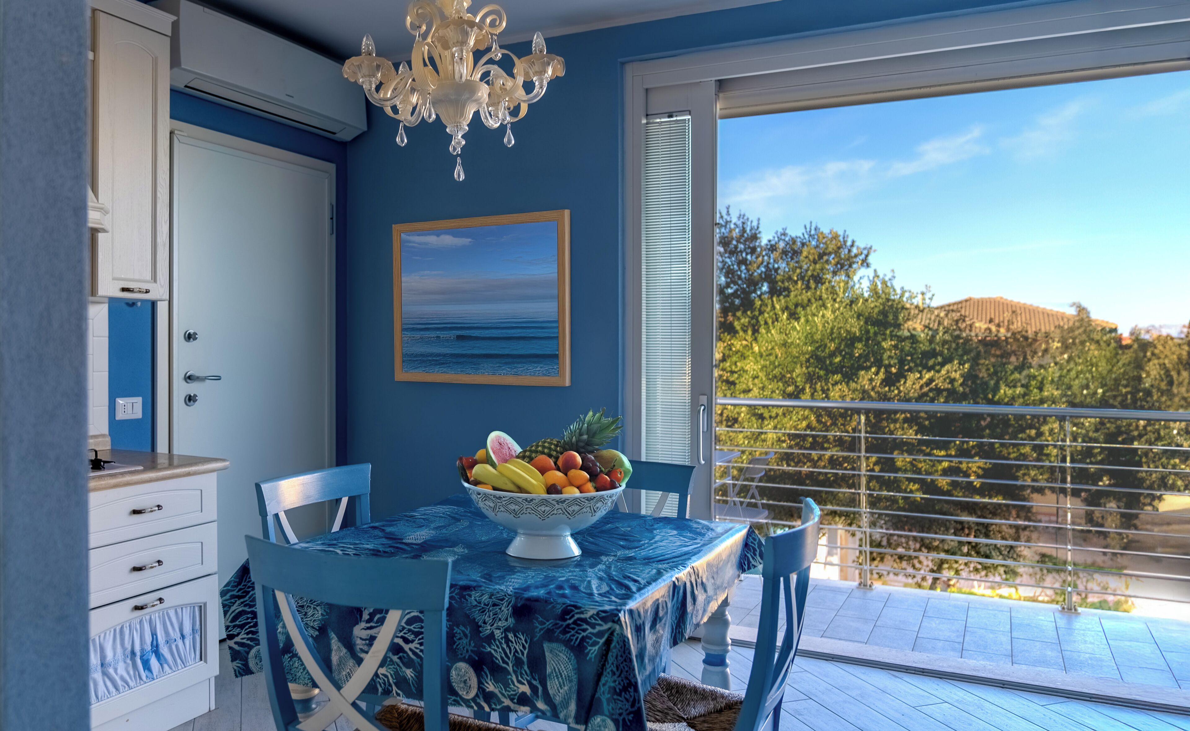 A room with a dining table set for two, a bowl of fruit, a framed picture of the sea, and a view of trees and hills through a balcony.