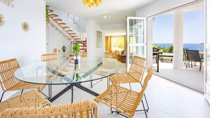 Ein modernes Interieur mit einem Glastisch, umgeben von Korbstühlen, einer Treppe und Blick auf das Meer.