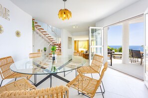 Ein modernes Interieur mit einem Glastisch, umgeben von Korbstühlen, einer Treppe und Blick auf das Meer.