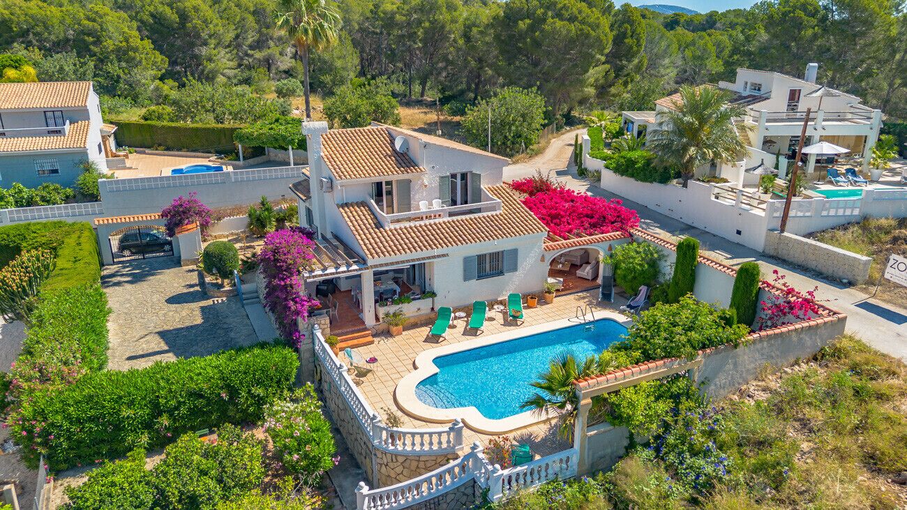 A two-story house with a swimming pool, surrounded by lush greenery and a white fence.
