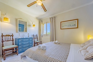 A bedroom with a bed, a dresser, a chair, a ceiling fan, a window with curtains, and a framed picture on the wall. - Villa Descanso - Plusholidays (Calpe / Calp)