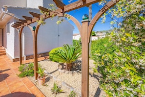 A garden with a wooden pergola, a palm tree, and flowering plants. - Villa Descanso - Plusholidays (Calpe / Calp)