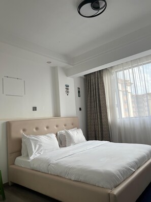 A hotel room with a large bed, a headboard, a window with curtains, and a wall-mounted light fixture. - Modern One-Bedroom serviced apartment/ 9th floor/ gym, lift, balcony, city view (Nairobi)