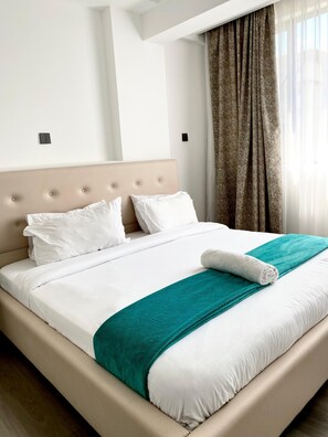 A neatly made bed with white linens and a teal throw blanket, a beige headboard, and a window with curtains. - Modern One-Bedroom serviced apartment/ 9th floor/ gym, lift, balcony, city view (Nairobi)