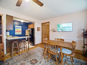 A dining area with a wooden table and chairs, a kitchen with blue walls and wooden cabinets, and a painting on the wall.