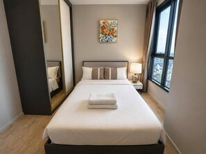 A hotel room with a large bed, a bedside table with a lamp, a mirror, and a window with a city view. - Sea View 2 Bed Room Best Location (Pattaya)