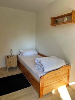 Ein Einzelbett mit Holzrahmen und weißer Bettwäsche, ein Nachttisch mit Lampe und ein kleines Regal über dem Bett.