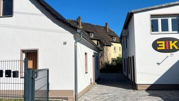 Ein schmaler gepflasterter Weg führt zu einem Gebäude mit weißer Fassade und einem schwarz-gelben Schild.
