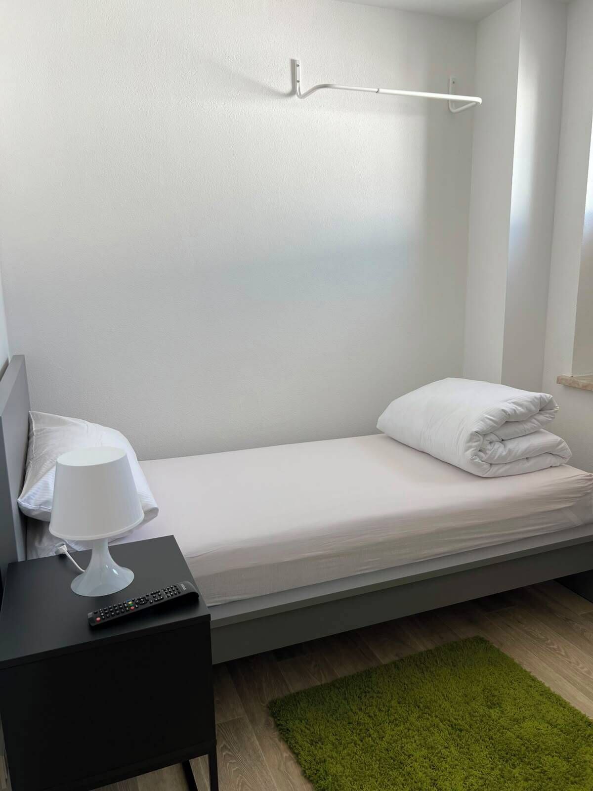 A single bed with a white mattress and pillows, a bedside table with a lamp and remote control, and a green rug on the floor.