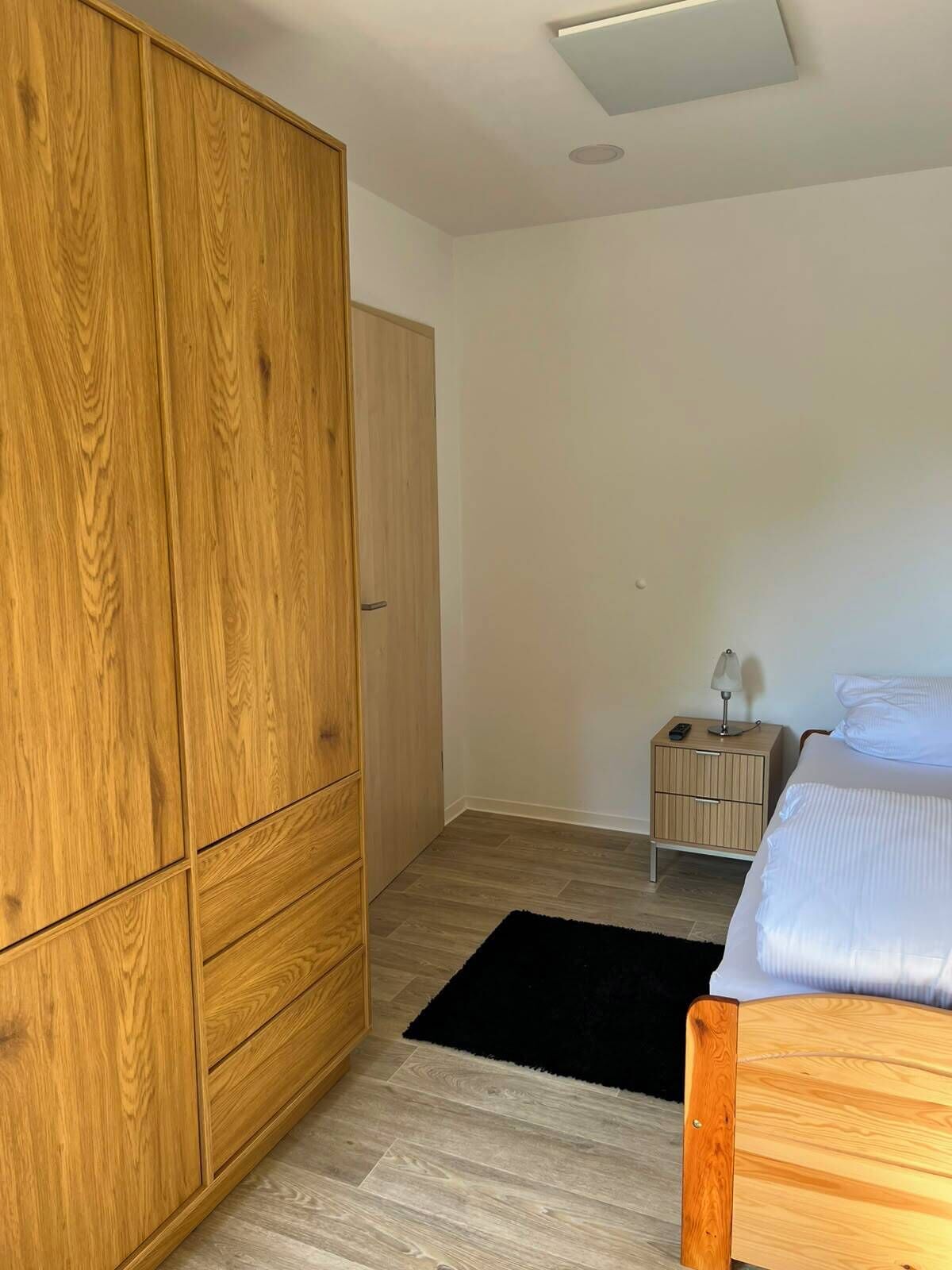 A bedroom with a wooden wardrobe, a bed, a nightstand, and a black rug.