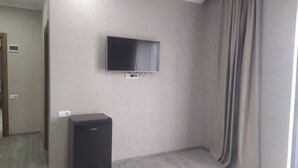 A room with a flat-screen TV mounted on the wall, a small black refrigerator, and a window with curtains. - ALPEDE (Zestafoni)