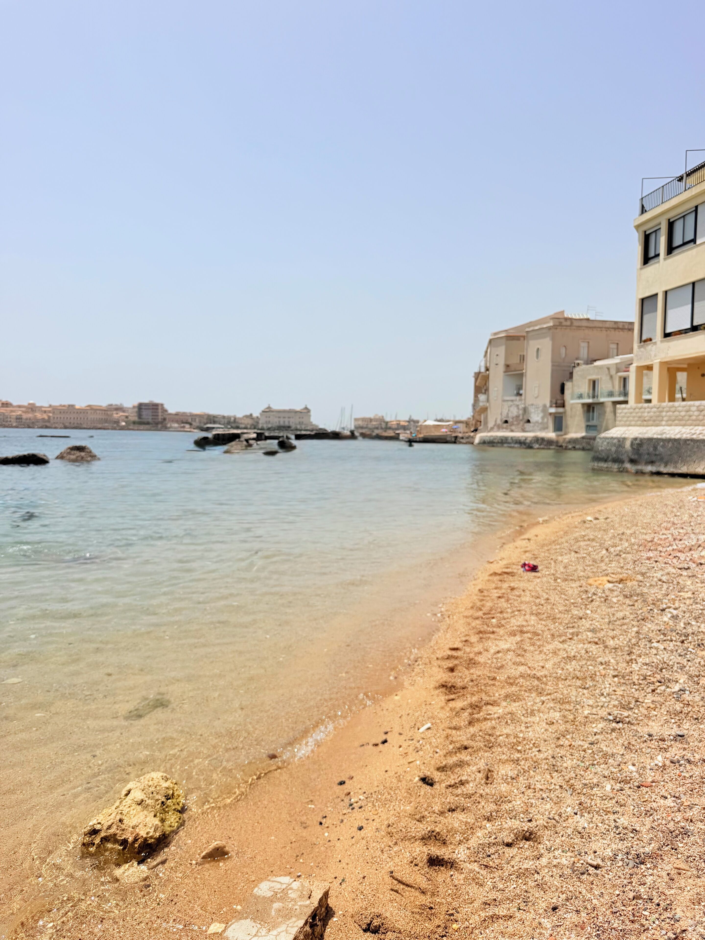A coastal area with clear water, a sandy beach, and buildings along the shore.