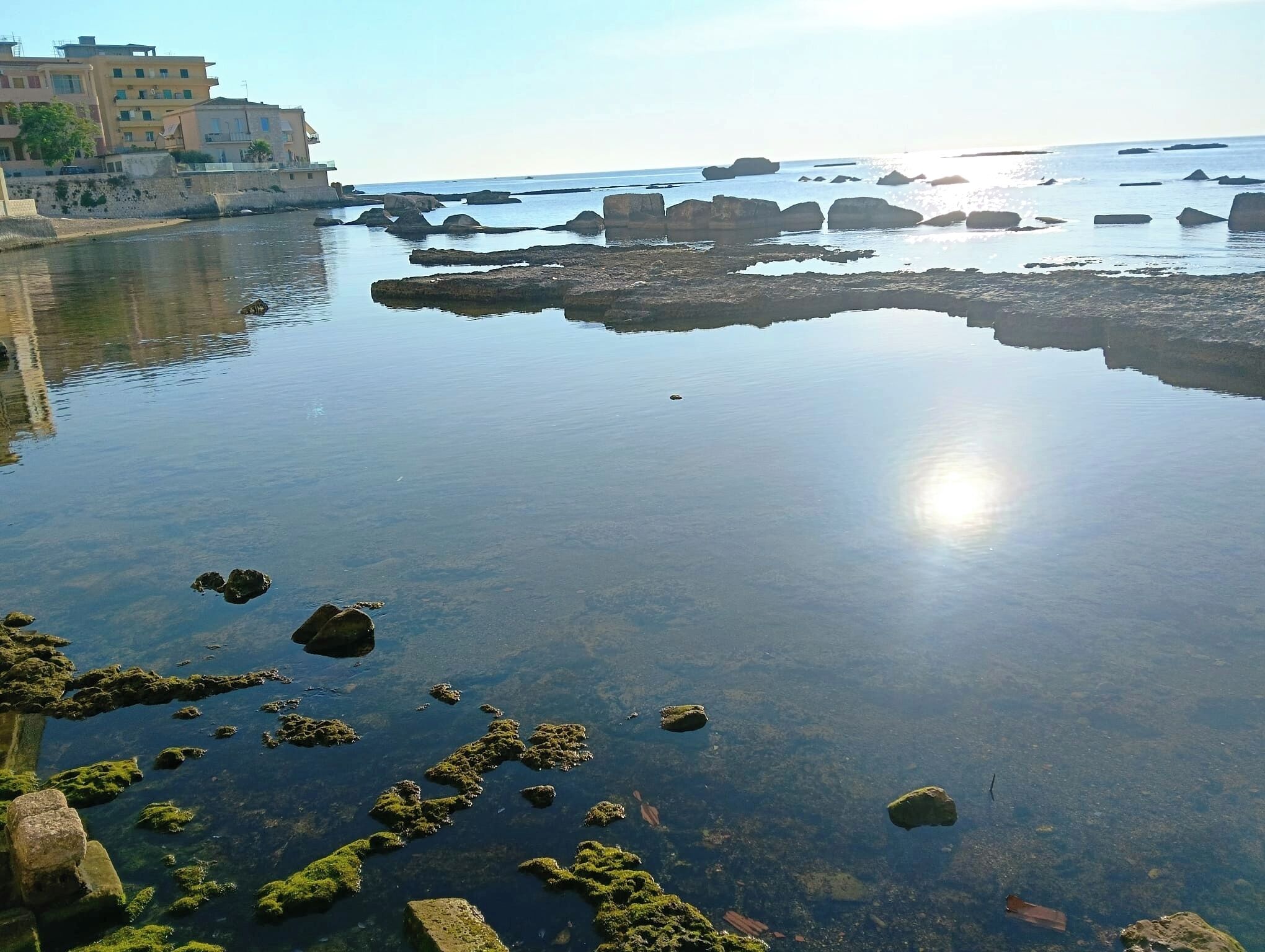 A coastal scene with clear water, rocks, and buildings in the background.