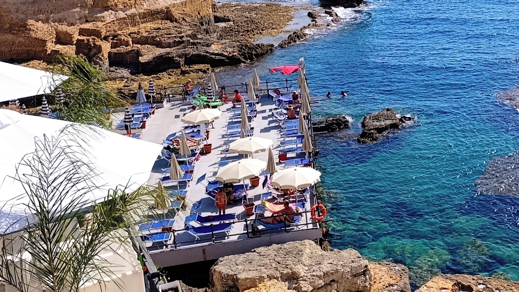 A coastal terrace with sun loungers and umbrellas, overlooking clear blue sea and rocky cliffs.