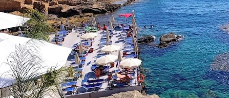 A coastal terrace with sun loungers and umbrellas, overlooking clear blue sea and rocky cliffs.