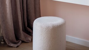 Quarto com uma mesa consola branca, uma planta em vaso, um banquinho redondo e uma janela com cortinas.
