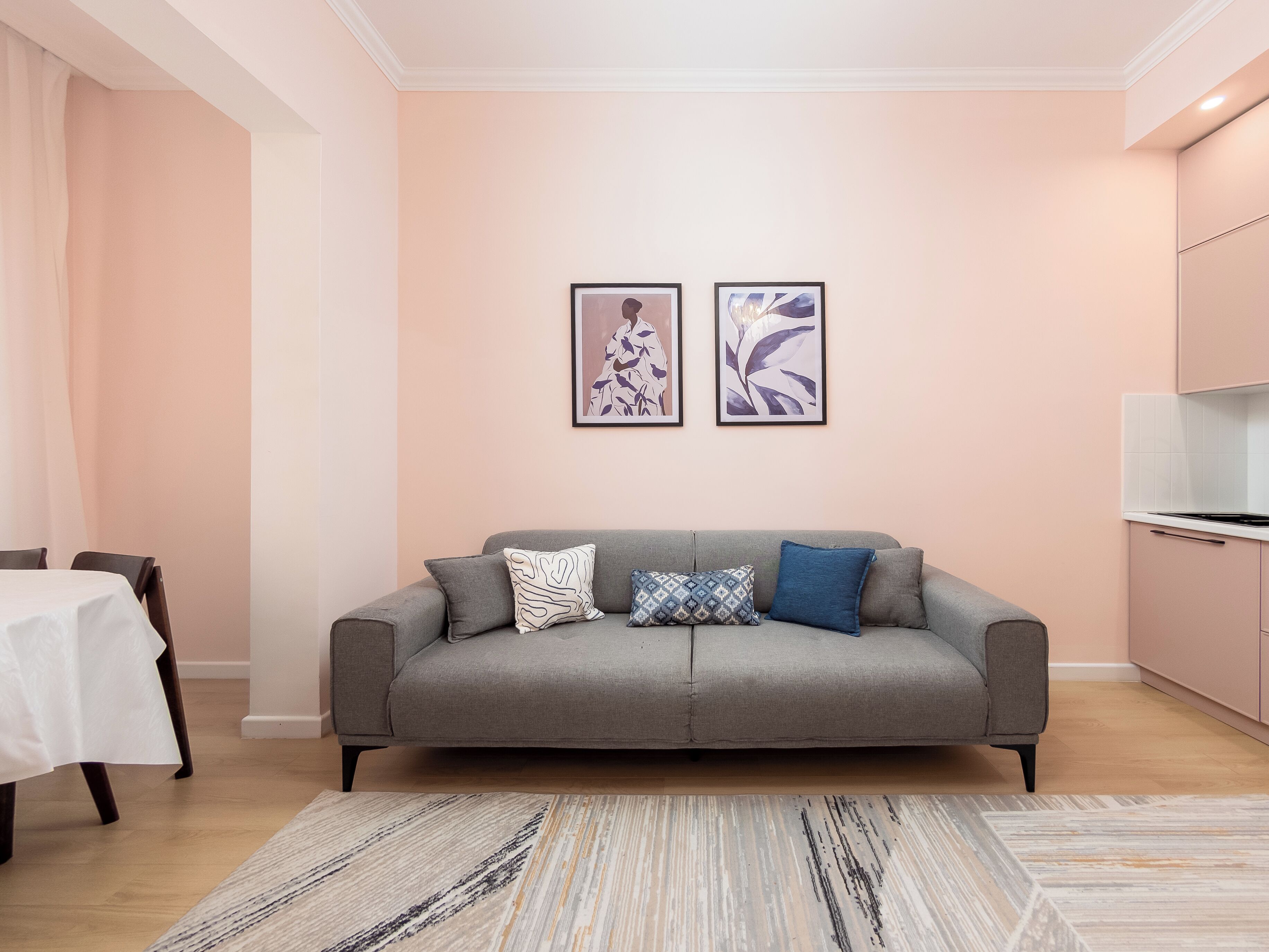 Een moderne woonkamer met een grijze bank, twee ingelijste kunstwerken, een kamerbreed tapijt en een keuken met roze kasten.