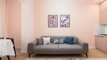 Een moderne woonkamer met een grijze bank, twee ingelijste kunstwerken, een kamerbreed tapijt en een keuken met roze kasten.