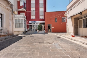 A narrow street with buildings on either side, a visible shop with a sign, and a small tree in the center.