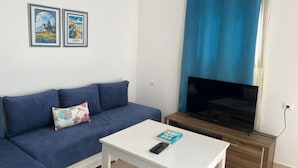 Una sala de estar con un sofá azul, una mesa de centro blanca, un televisor de pantalla plana y dos cuadros en la pared.