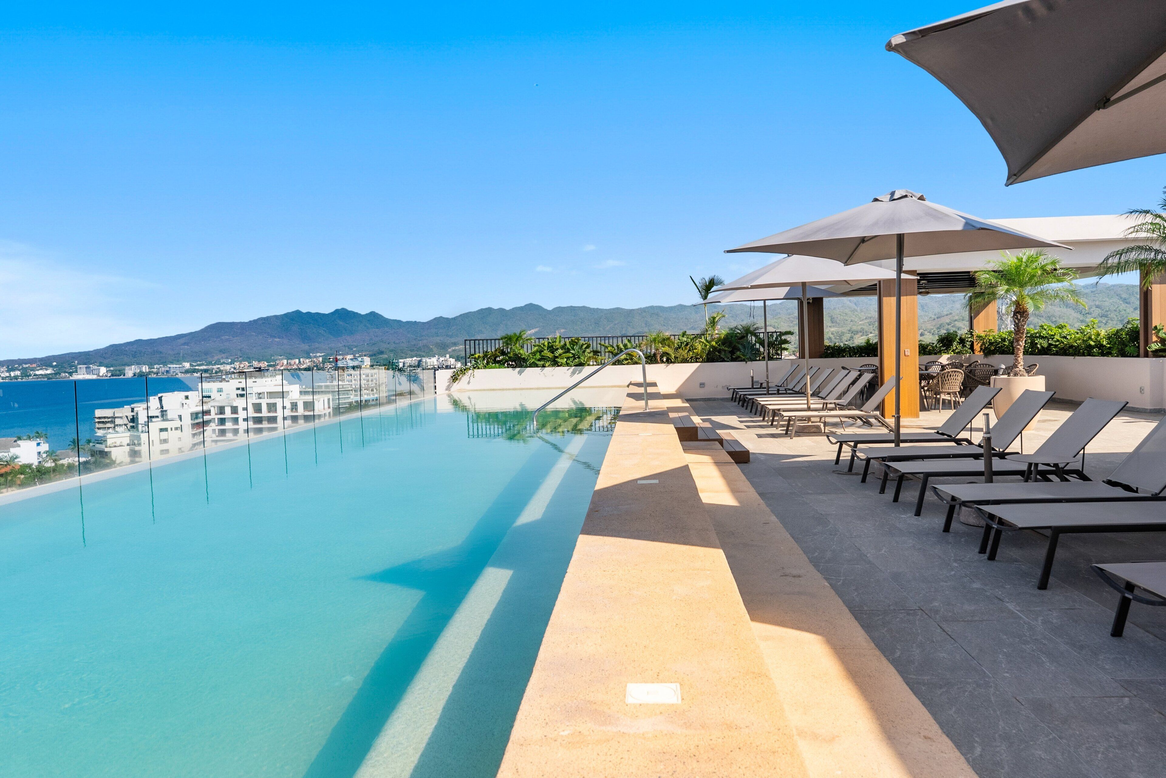 Ein Poolbereich mit Liegestühlen und Sonnenschirmen, mit Blick auf ein Gewässer und eine ferne Skyline.