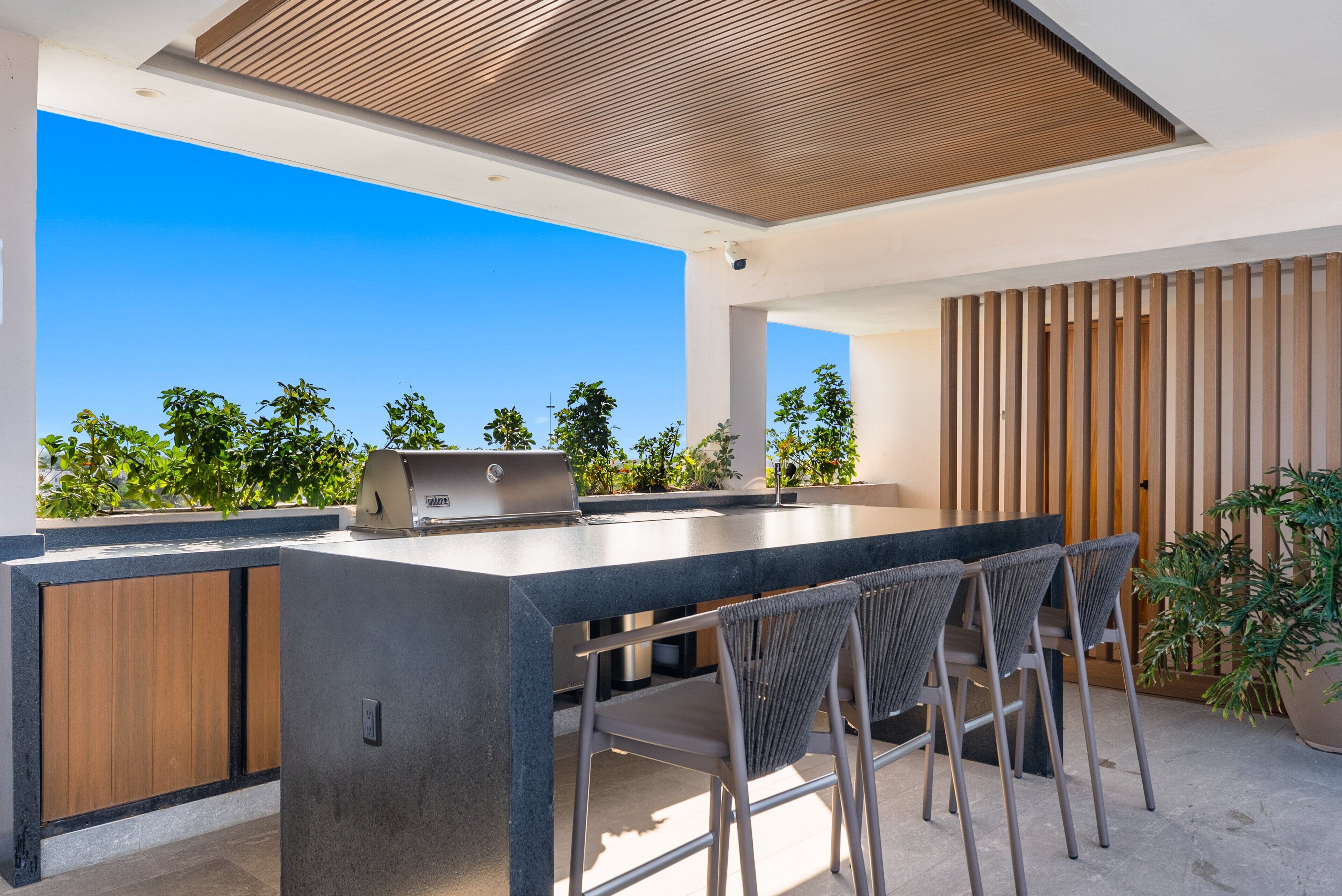 Una cocina al aire libre moderna con una parrilla integrada, asientos tipo bar y vistas al cielo y la vegetación.