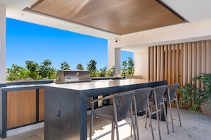 A modern outdoor kitchen with a built-in grill, bar seating, and a view of the sky and greenery. - 3BR condo near the ocean in Bucerías with pool, WiFi & full kitchen (Bucerias)