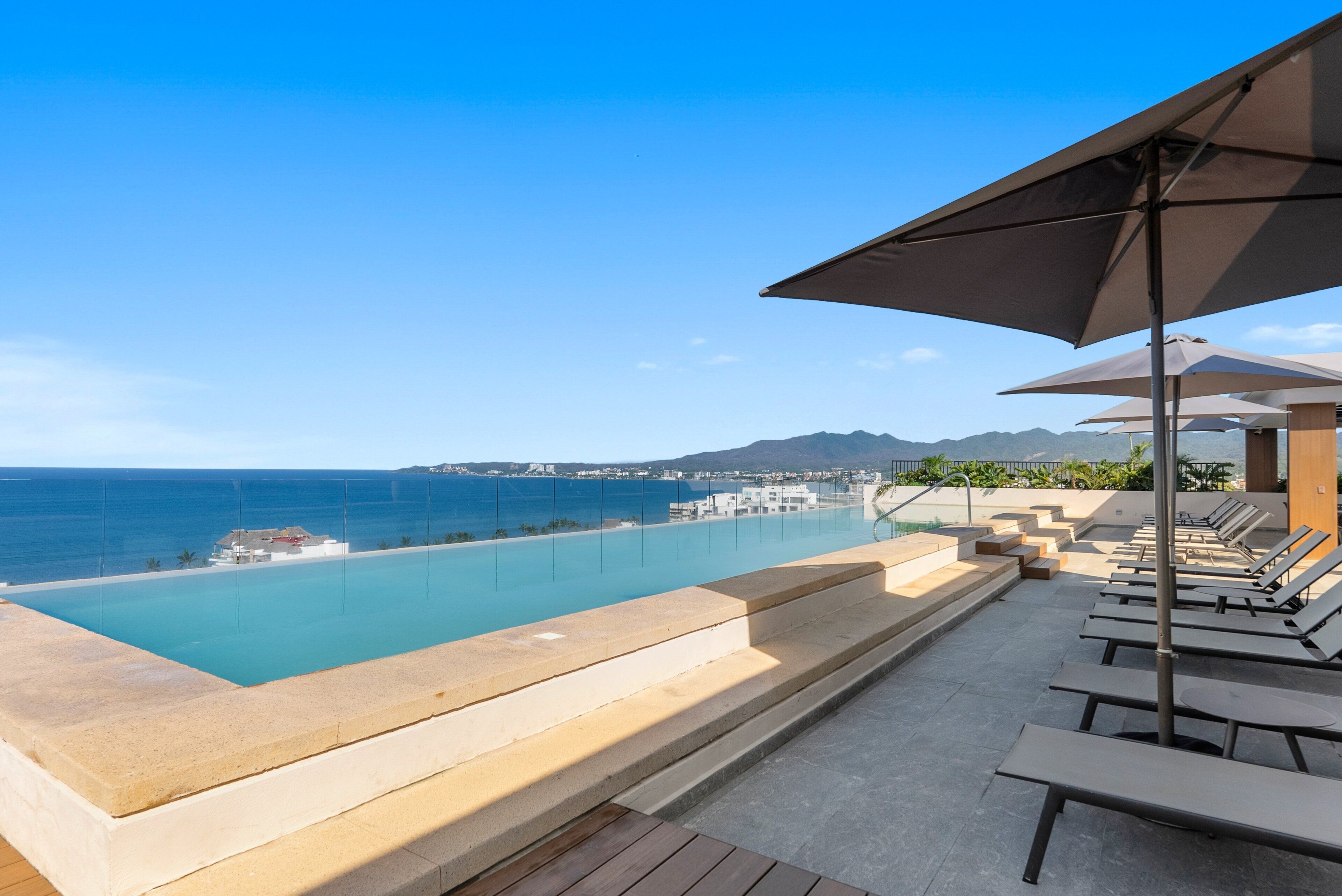An infinity pool with a view of the ocean and distant mountains, surrounded by lounge chairs and umbrellas.