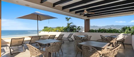 Un patio al aire libre con mesas y sillas, un ventilador de techo y vista al océano.