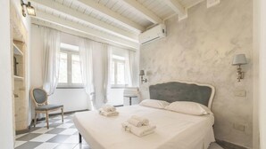 A hotel room with a bed, a chair, a window with curtains, a wall-mounted lamp, and a checkered floor. - Estay - The Corner House Cagliari Room 1 (Cagliari)