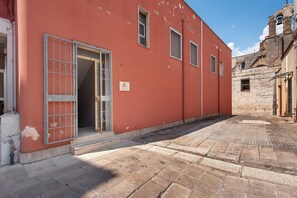 A red building with a door and windows, a stone-paved courtyard, and a bell tower in the background.