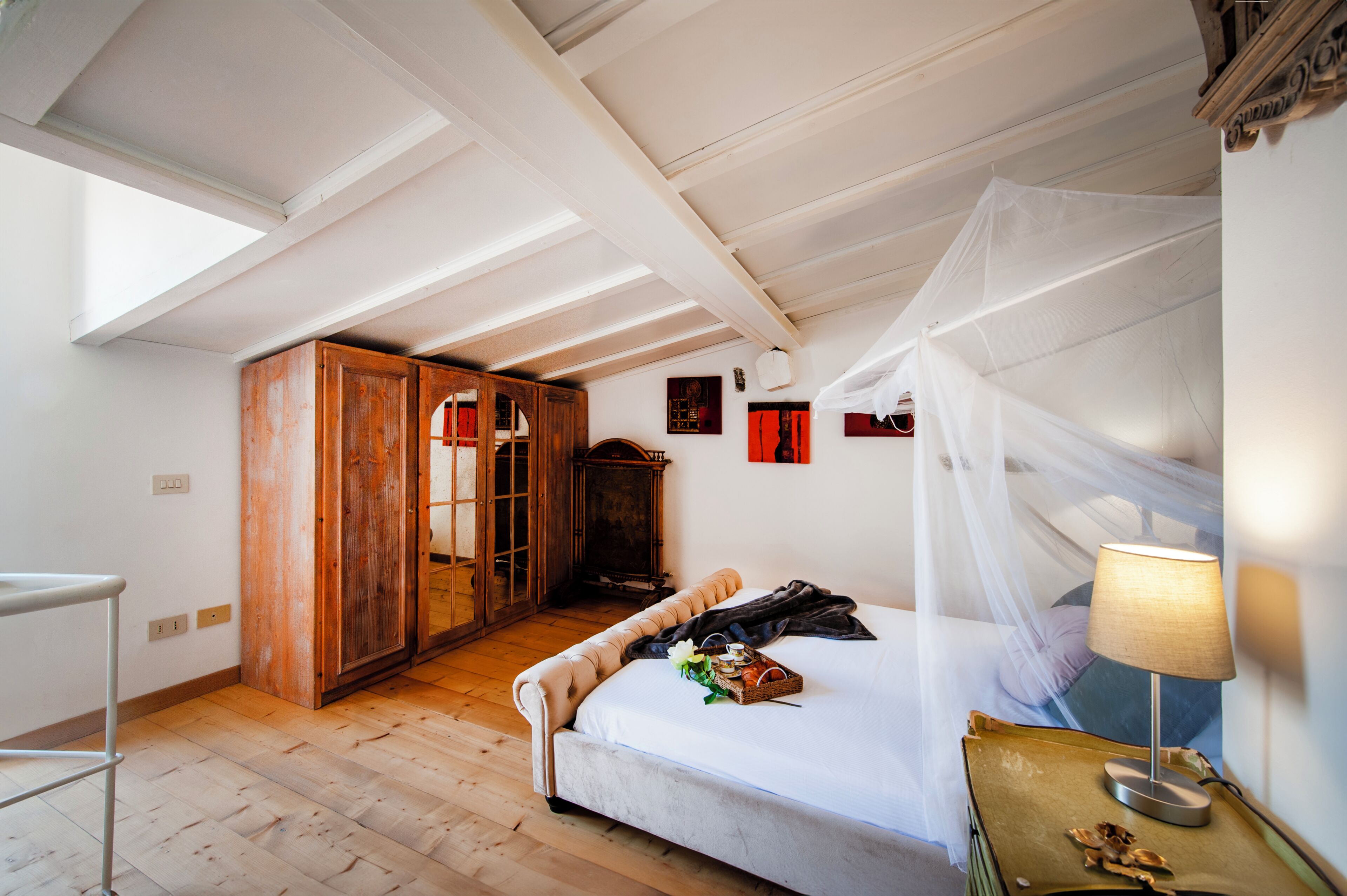 Quarto com cama, guarda-roupa de madeira, uma mesinha com abajur e janela com dossel branco.