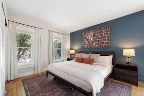 Une chambre à coucher avec un grand lit, deux tables de chevet, une œuvre d’art accrochée au mur et une vue par la fenêtre.