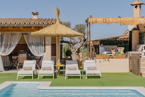 Ein Poolbereich mit Liegestühlen, einem Reetdach-Sonnenschirm und einer Essgelegenheit mit Gartenmöbeln.