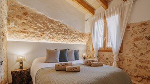 Een slaapkamer met een stenen muur, een bed met kussens, een nachtkastje met een lamp en een raam met gordijnen.