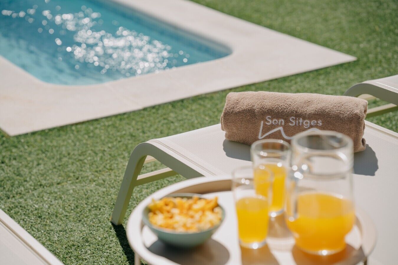 Une scène au bord de la piscine avec une serviette sur laquelle est écrit « Sons et images », un bol de céréales et des verres de jus d’orange.