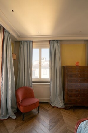 Ein Zimmer mit Fenster, Stuhl, Kommode und Lampe.