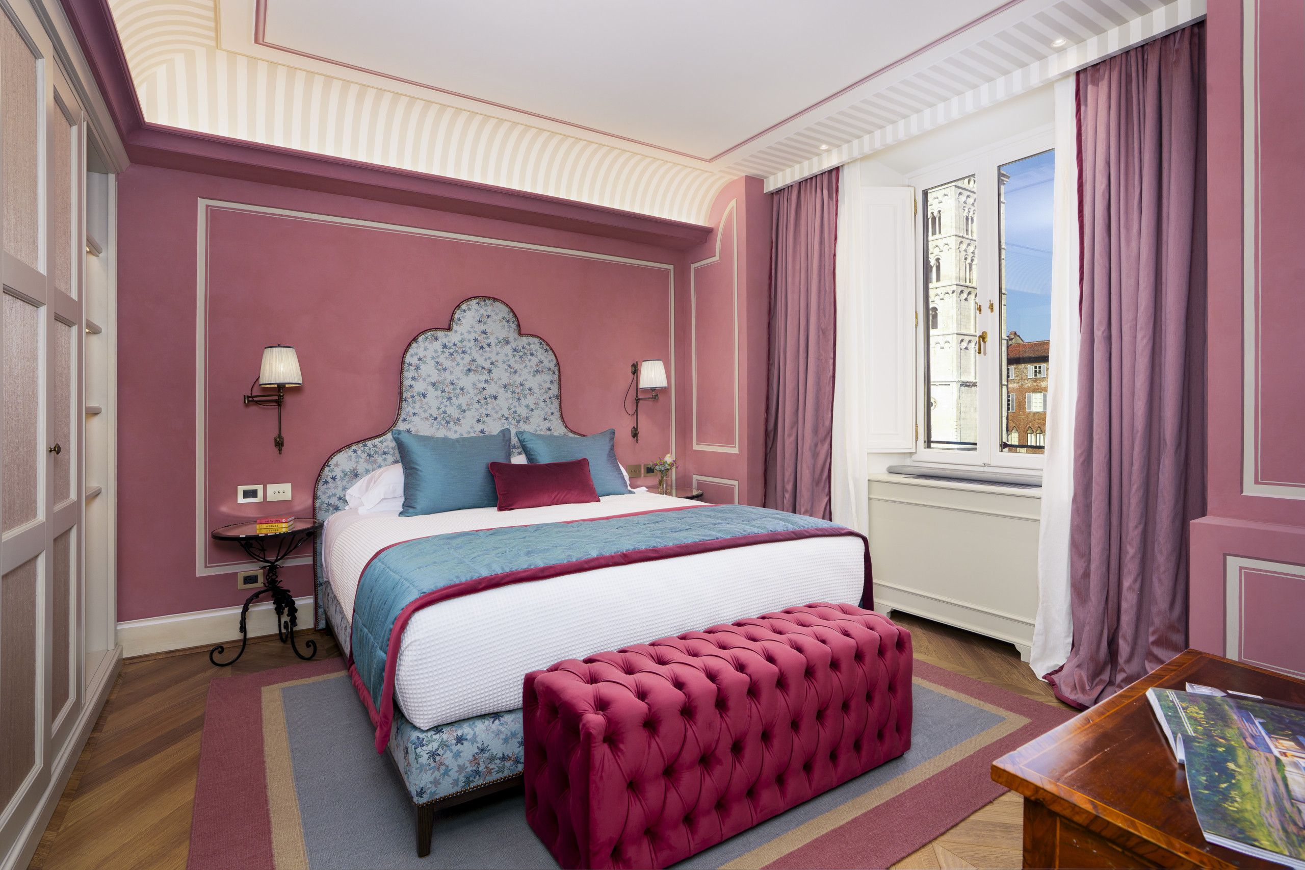 Un dormitorio con una cama grande, una mesita de noche, una lámpara, un ventanal con cortinas y vistas a un edificio.