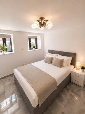 A bedroom with a bed, bedside tables, a chandelier, and windows with plants. - Modern & Elegant Apartment | King Bed (Bucharest)