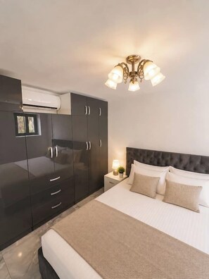Quarto com cama grande, guarda-roupa escuro, mesa de cabeceira com abajur e luminária no teto.
