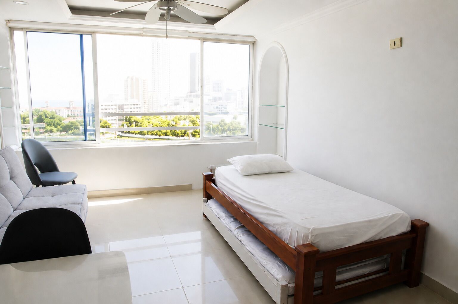 Une chambre à coucher comprenant un lit, une chaise, une fenêtre donnant sur des bâtiments et de la verdure, ainsi qu’un ventilateur de plafond.