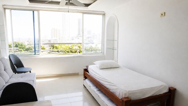 Un dormitorio con una cama, una silla, un ventanal con vistas a edificios y follaje, y un ventilador de techo.