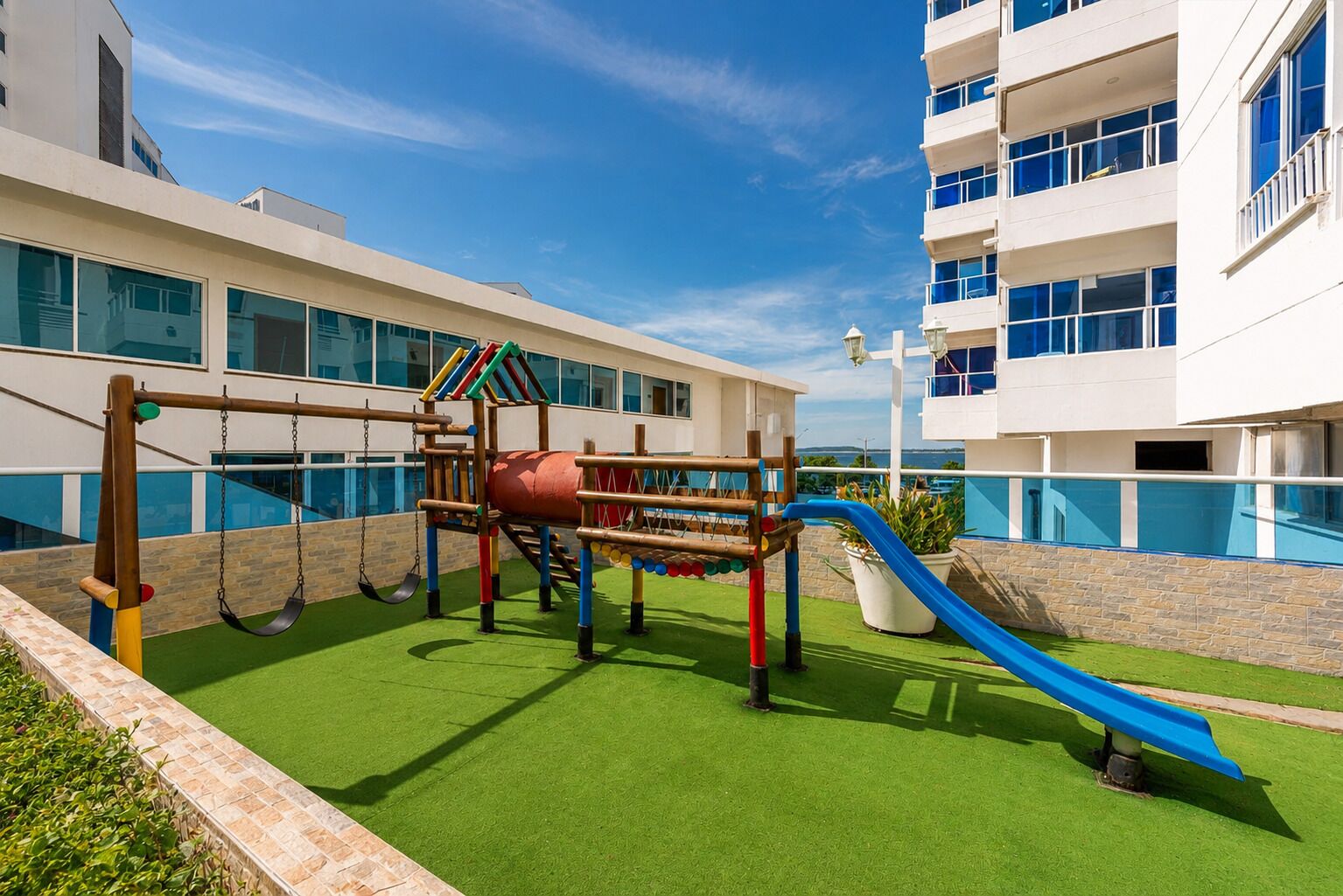 Un parc infantil amb tobogan i cadira de balanceig, tot envoltat d'un edifici amb grans finestres i una piscina.
