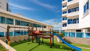 Un parque infantil con tobogán y columpios, rodeado por un edificio con ventanas amplias y una piscina.