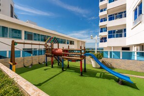 A playground with a slide and swings, surrounded by a building with large windows and a pool. - Encantador Apartamento Frente a la Playa (Cartagena de Indias)