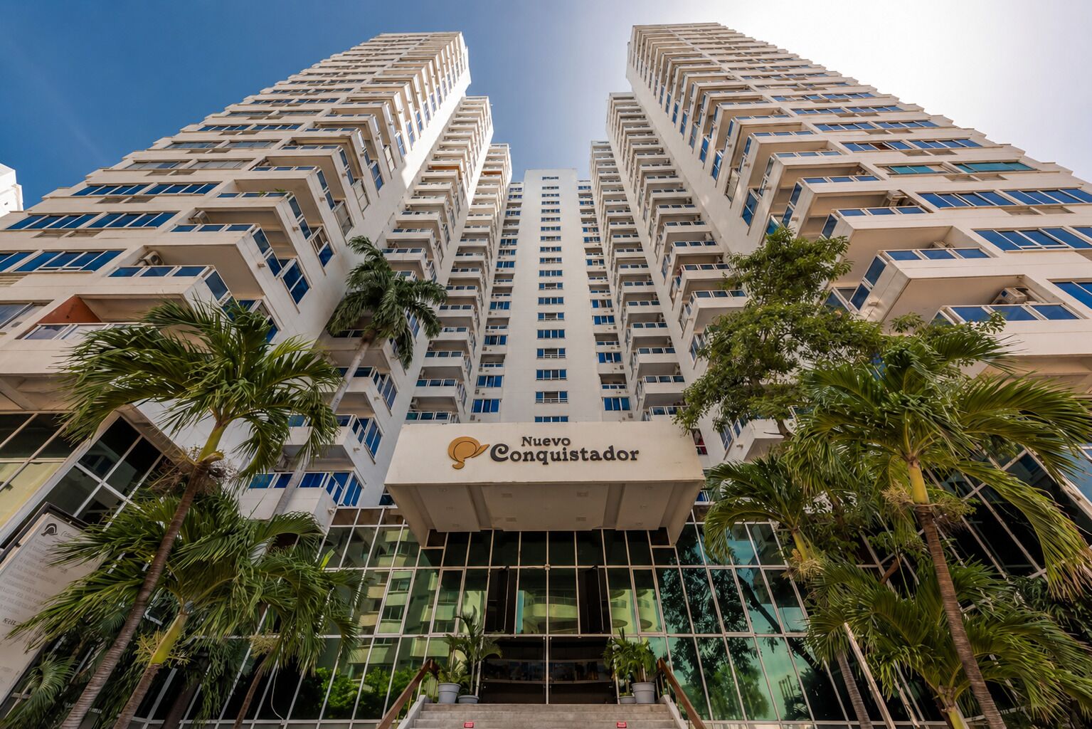 A high-rise hotel building with multiple floors, a prominent entrance, and surrounding palm trees.