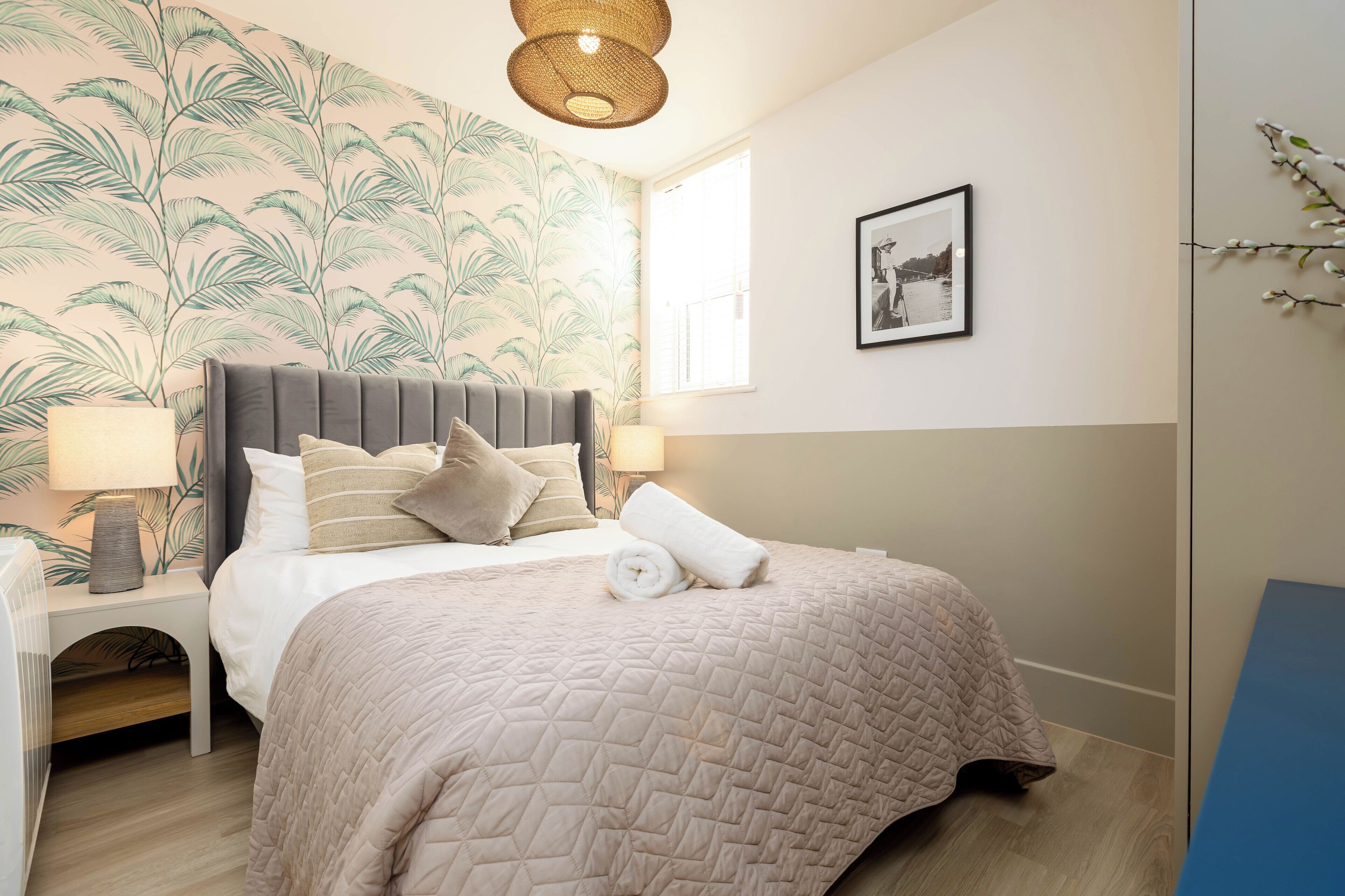 A bedroom with a bed, a nightstand, a lamp, a wall-mounted picture, and a window.