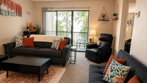 A living room with a sofa, a coffee table, and a chair. There is a window with a view of trees.