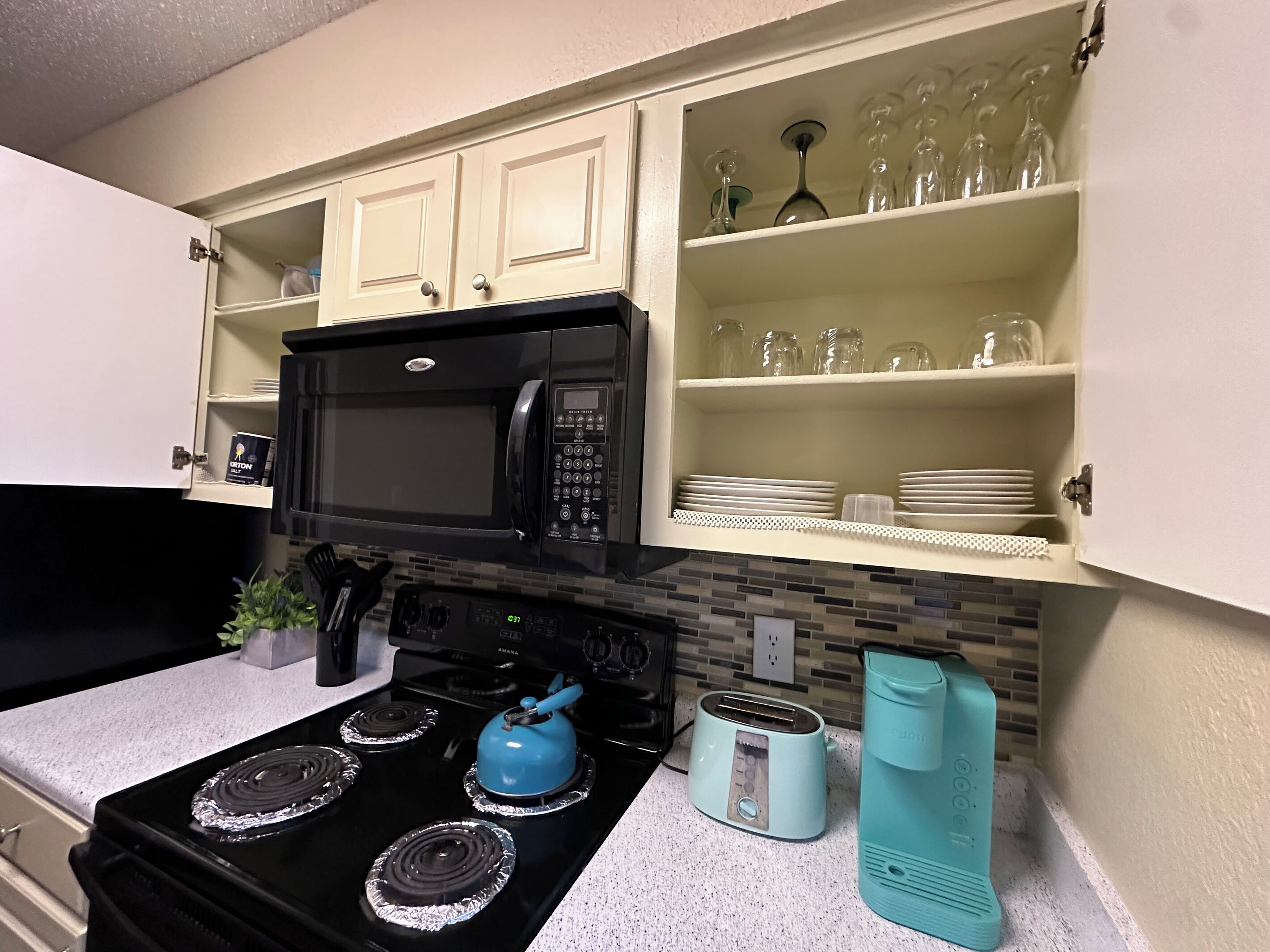 A kitchen with a black stove, a microwave, and a toaster. There are cabinets above the stove and a shelf with glassware.