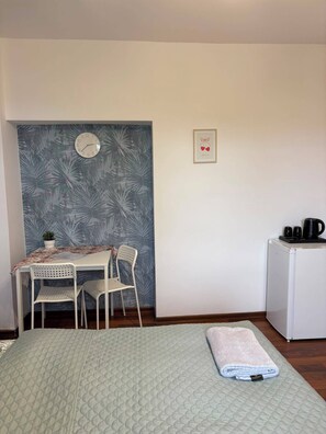 A room with a bed, a small table with chairs, a clock, a framed picture, and a refrigerator.